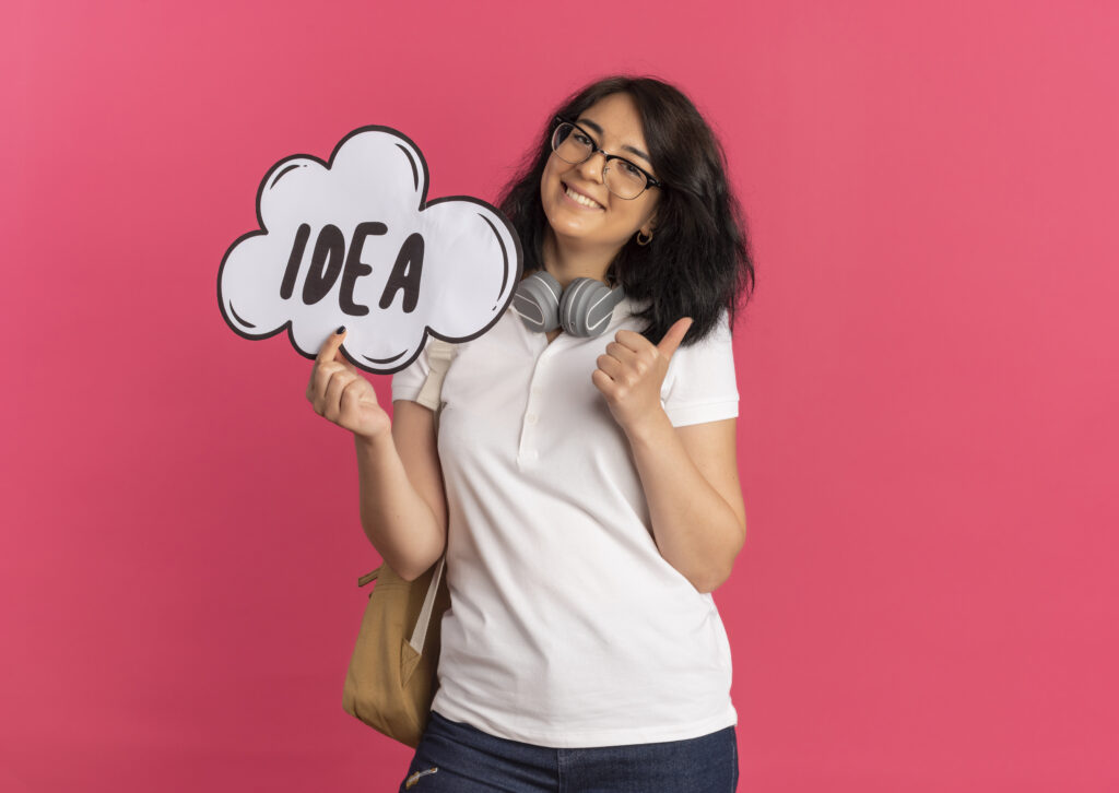 young-smiling-pretty-caucasian-schoolgirl-wearing-glasses-back-bag-headphones-neck-holds-idea-mark-thumbs-up-pink-with-copy-space-1024x726 Você é o Seu Maior Projeto: Como Construir a Si Mesmo com Propósito, Consistência e Autoconsciência